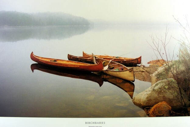 Full color photography page from a book, showing three replica birchbark canoes gathered near a section of the rocky shore that's further into the water. The water is as smooth as glass. Fog obscures the background.