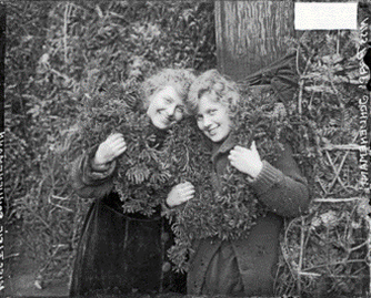 Hazel and Pearl Schunemann smile at the camera, wreaths around their necks and christmas trees behind them